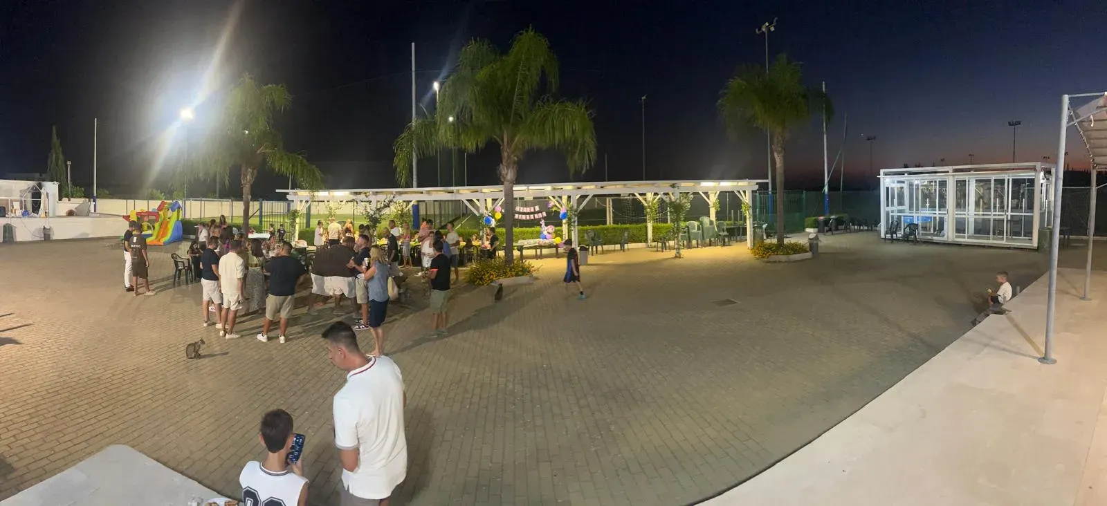 Vista panoramica serale di una festa privata nel piazzale del Champions Park, con ospiti, palme e pergolato illuminato